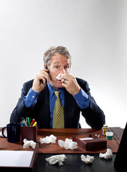 A middle-aged man sits at a desk, talking on the phone and trying to blow his nose