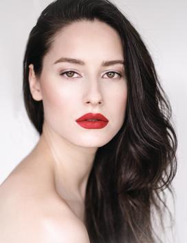 head and shoulder beauty shot of a Caucasian female with long brunette hair. glowing skin and light shimmery eye make up and a matt red lip.