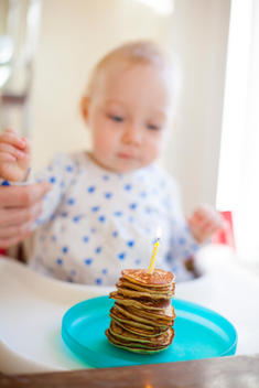 A tiny stack of pancakes with one lit birthday candle sit in front of a one year old baby.