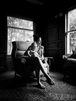 black and white image of thirty something woman holding a baby while sitting in vintage chair by large window.