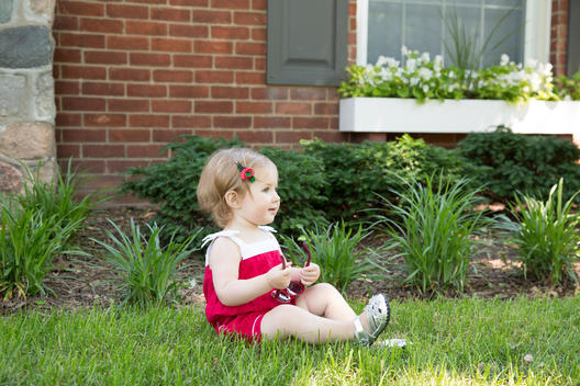 Baby girl sitting in front yard of house, holding sunglasses in lap.