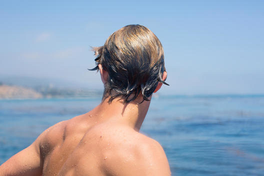 Portrait of back of the head of early twenties brown haired man with no shirt on and wet hair with the Pacific Ocean in the background.