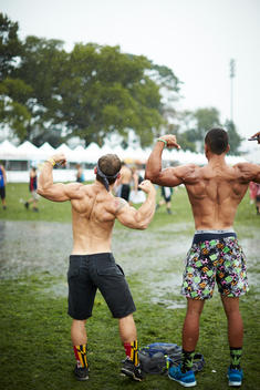 Two shirtless men flexing from behind after a rainstorm at a music festival
