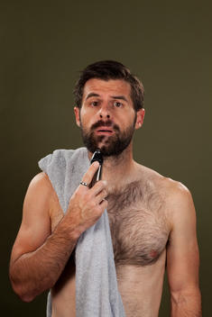 A man with a towel over his shoulder holds up an electric razor to his beard wear a swath of hair has been shaved off