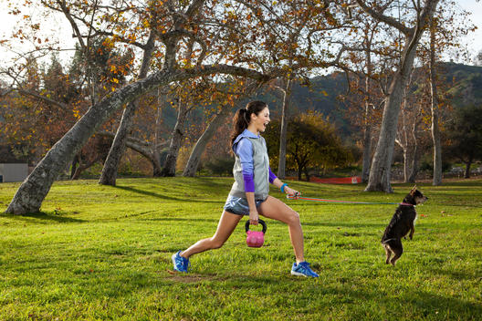A woman in workout gear stretches in a park while trying to hold onto the leash of her jumping dog