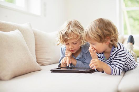 Two boys lying on fronts side by side on sofa using digital tablet