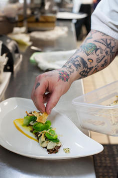 chef with tattooed arm garnishing dish in kitchen