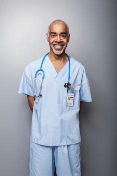 Male, African American Doctor with grey hair, wearing medical scrubs on grey background.