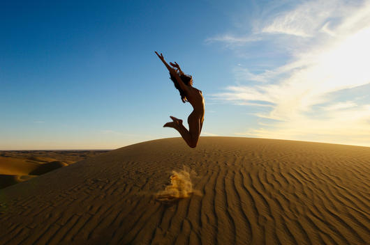 Nude athletic young woman jumping up in the air on a sand dune against a blue sky