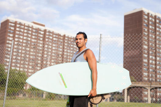 Portrait of surfer muscular, athletic Matt Hernandez with large wings tattoo on chest holding a surfboard with projects, government housing behind at Far Rockaway Beach, Queens