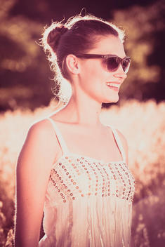 young woman with sunglasses standing at summer in a cornfield, wearing a light dress and smiling, back light and lens flare