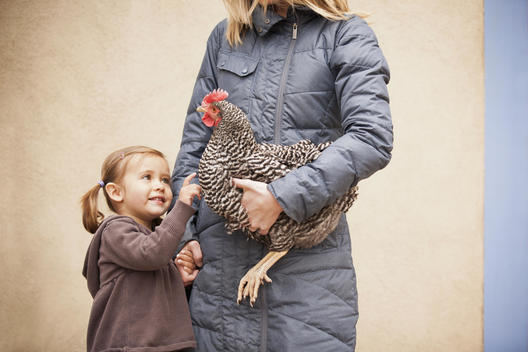 A woman in a grey coat holding a black and white chicken with a red coxcomb under one arm. A young girl beside her holding her other hand