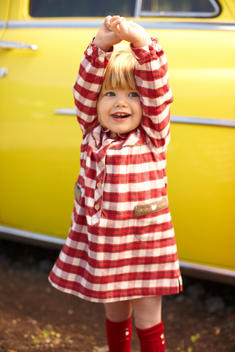 Toddler girl in checked red dress in front of yellow car, holding her hands over her head.