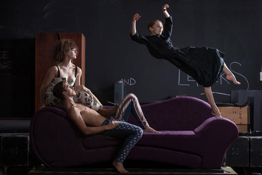 A beautiful woman falling off a couch towards two of her friends, suspended above them.