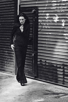 Caucasian woman with long dark hair in stripe trouser suit, walking pass a garage gate, looking down