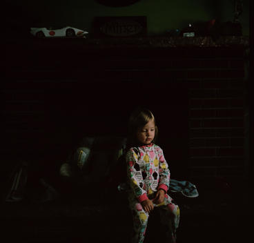 A young blonde girl wearing cupcake pajamas sits on the edge of the fireplace in the dark morning light in the her family\'s suburban home.