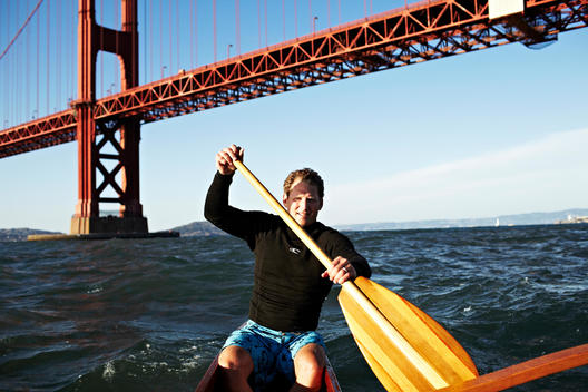 Young man paddling a wooden outrigger canoe under the Golden Gate Bridge.