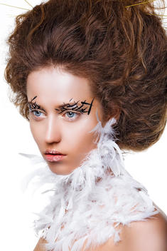 Shot of the model with volume hair and unusual makeup, like a bird. There is a plumage on her neck.