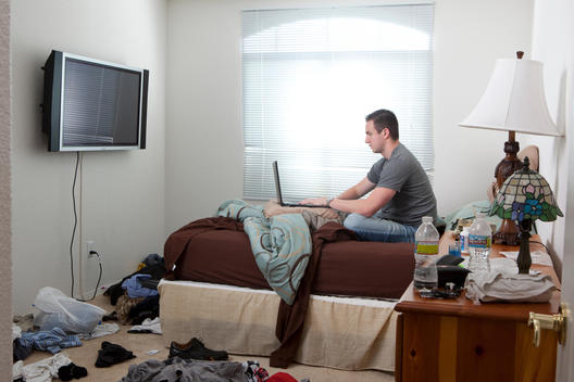 In a messy bedroom, a man sits on his bed playing on his laptop computer