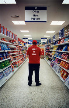 Back Of A Supermarket Worker Wearing A Red Shirt That Says \'Here To Help\', Standing In The Supermarket Isle