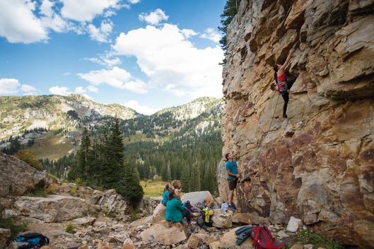 A woman rock climbing up a steep rock wall with four other people watching and belaying in a side canyon up Little Cottonwood Canyon near Salt Lake City, Utah