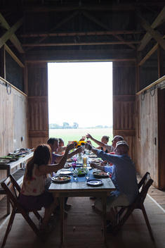 A group of people including Tom Colicchio raising glasses together at a dinner table inside a barn