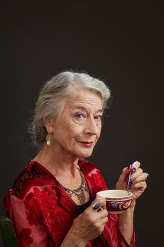 A formally dressed old woman holds a teacup in one hand while stirring it with a spoon in the other