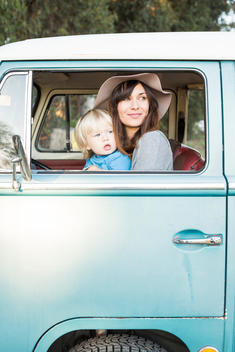 2 year old blonde boy in blue shirt and young mom with straw hat looking out vintage van.
