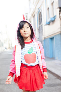 6 year old Asian girl looking at the camera and wearing a pink dinosaur hoodie on the street in San Francisco, CA.