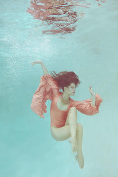 Woman underwater in pink leotard and long sleeves crossing her legs and stretching her arms