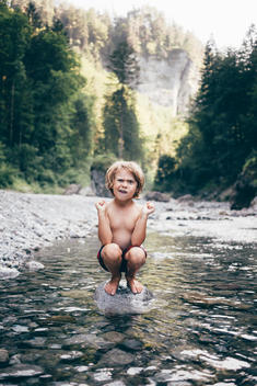 6 year old blonde boy in swim shorts sitting on rock in clear water creek in Austrian alps.