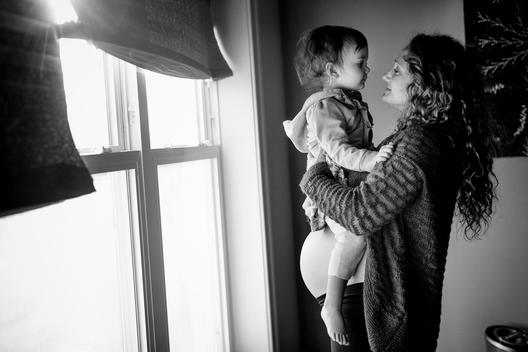 Black and white image of pregnant mom and young daughter embracing by window light