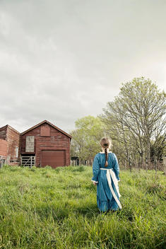 A young girl with her long brown hair in a plait/braid, dressed in an olden day style blue dress with cream bow, stands in long green grass facing towards a rustic barn. The sky is filled with grey and white clouds. Trees and a wire fence run along the le