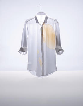 A still life image of a mans shirt with a big coffee stain against a white background.