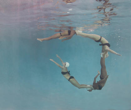 Four synchronized swimmers underwater in white caps and blue bathing suits in the process of forming a dolphin circle