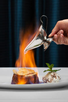 Hand Pouring Alcohol In Silver Pitcher Onto Flaming Bananas Foster Flambé Dessert.
