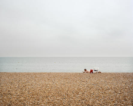 A man sunbathing on Brighton Beach whilst reading a book. The English Channel can be seen in the distance.