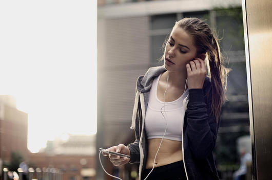 A young female runner dressed in sports clothing in urban surroundings listening to music through earphones from a portable music player