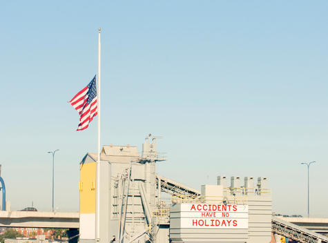 US flag flying at half mast over industrial buildings displaying warn signs about health and safety at work