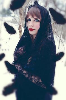 Photograph of a young caucasian woman with blonde hair, striking blue eyes, and red lipstick. She is covered with a black lace shawl and standing in a wooded area that is blanketed in snow. She is smiling at the camera with black feathers surounding her.