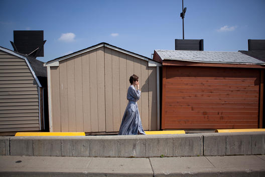 A woman smoking a cigarette while walking past a selection of garden sheds, Queens, NY.