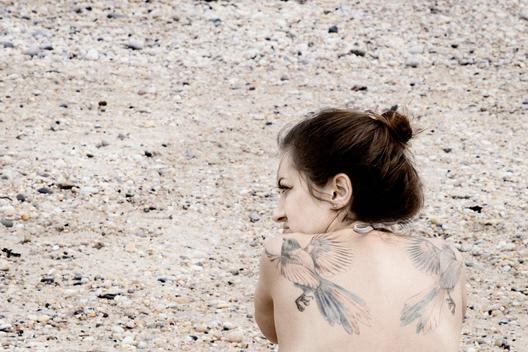 Young Woman With Bird Tattoos Sitting On The Beach In Early Spring