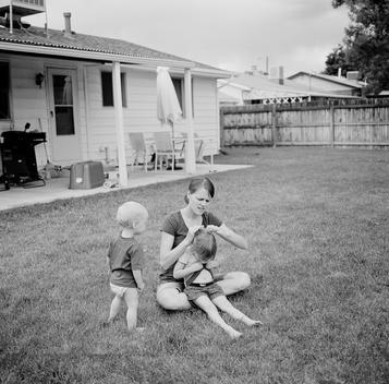 Kim, a young mother of two children, puts her daughter Jane\'s hair in a ponytail while sitting on her lap as her baby son Grant attentively watches on a sunny summer afternoon in their suburban backyard. Denver, Colorado