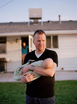 A middle-aged father lovingly cradles his newborn son in the backyard of his family's suburban home in late afternoon setting sun with his wife standing in the home doorway in background. Denver, Colorado