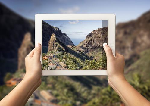 Hands of young woman holding up digital tablet with view of Masca Valley in front of view of Masca Valley, Tenerife, Spain