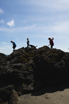 three boys and a dog climbing over a rock on a beach, silhouetted against the sky