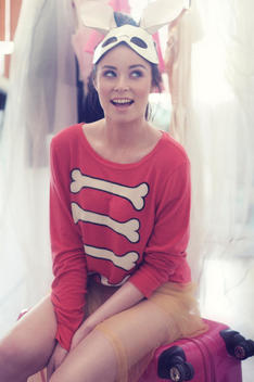 Caucasian teen girl with dark hair in a bun.Smilling looking up with a mask with bunny ears on her head,wearing a red sweat shirt sitting on a pink suitcase