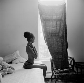 Dramatic black and white portrait of Lisa, a beautiful semi-nude black teen model topless in leopard panties sitting alone on bed near window in an empty bedroom. Brooklyn, New York