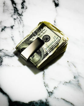 A wad 100 dollar bills in cash with a money clip on a marble surface