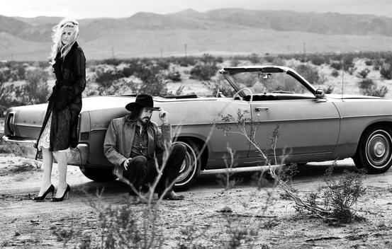 A model couple out in the desert lean against their old convertible Cadillac.